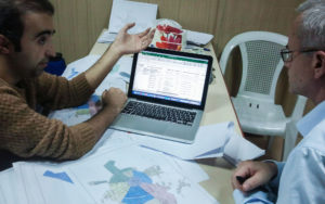 Two persons working together on a laptop, with maps on the table.